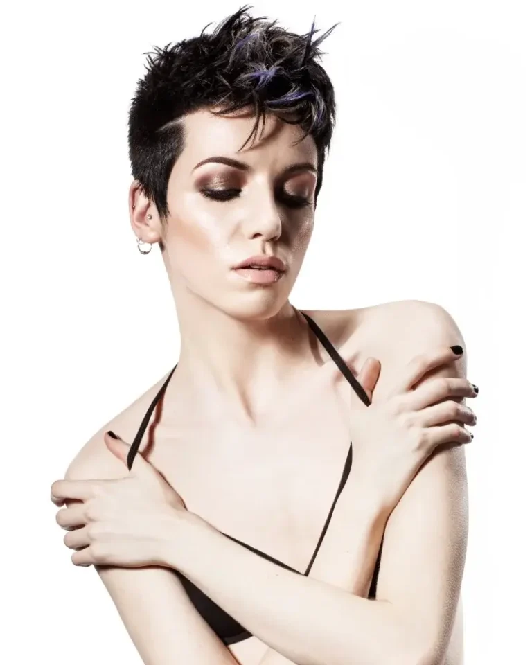 Model with a cropped, textured dark pixie cut featuring subtle blue highlights and shaved sides, posing with arms crossed