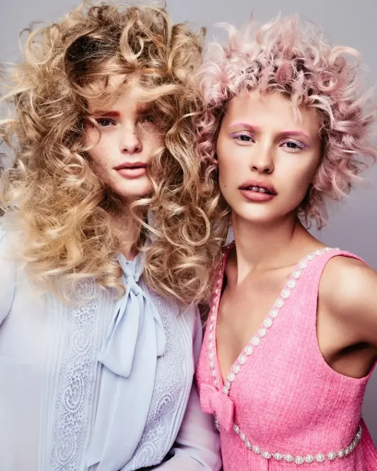 Two models side by side featuring voluminous blond curls and pastel pink textured afro-style curls