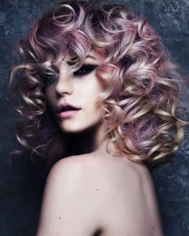 Model with voluminous, curly platinum blond hair highlighted with pastel pink and violet tones looking over her bare shoulder