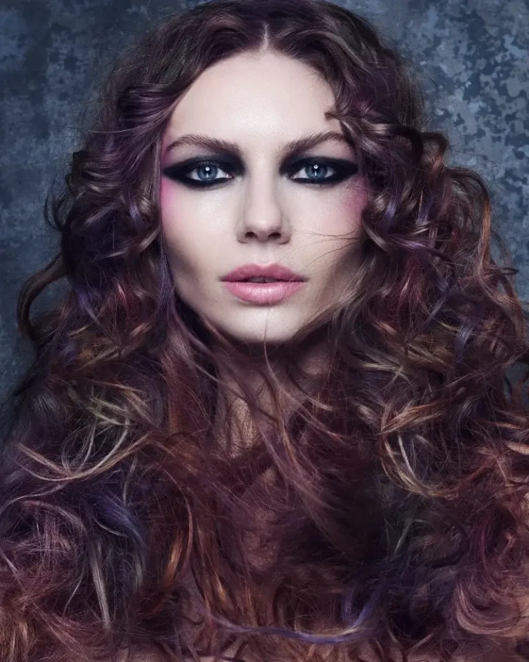 Model with long, rich dark curls layered with subtle plum and burgundy tones facing forward with dramatic eye makeup