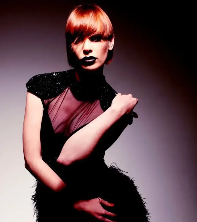 Editorial portrait of a woman with a precision-cut copper red crop featuring a smooth side-swept fringe, dressed in a black sheer embellished top